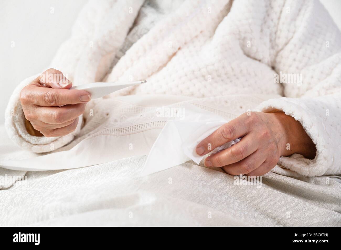 Coronavirus. Sick senior woman holding tissue and thermometer sit on bed, upset old mature woman caught cold got coronavirus flu influenza symptoms at home alone . Stock Photo