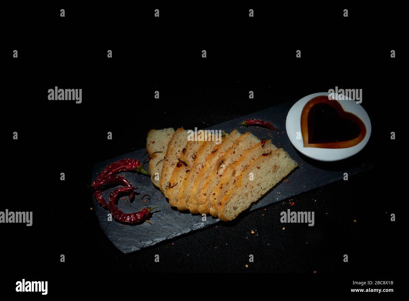 Display of homemade Italian style Focaccia bread with red chillies.  Sliced bread on black slate with balsamic vinegar in heart shape dish a Stock Photo