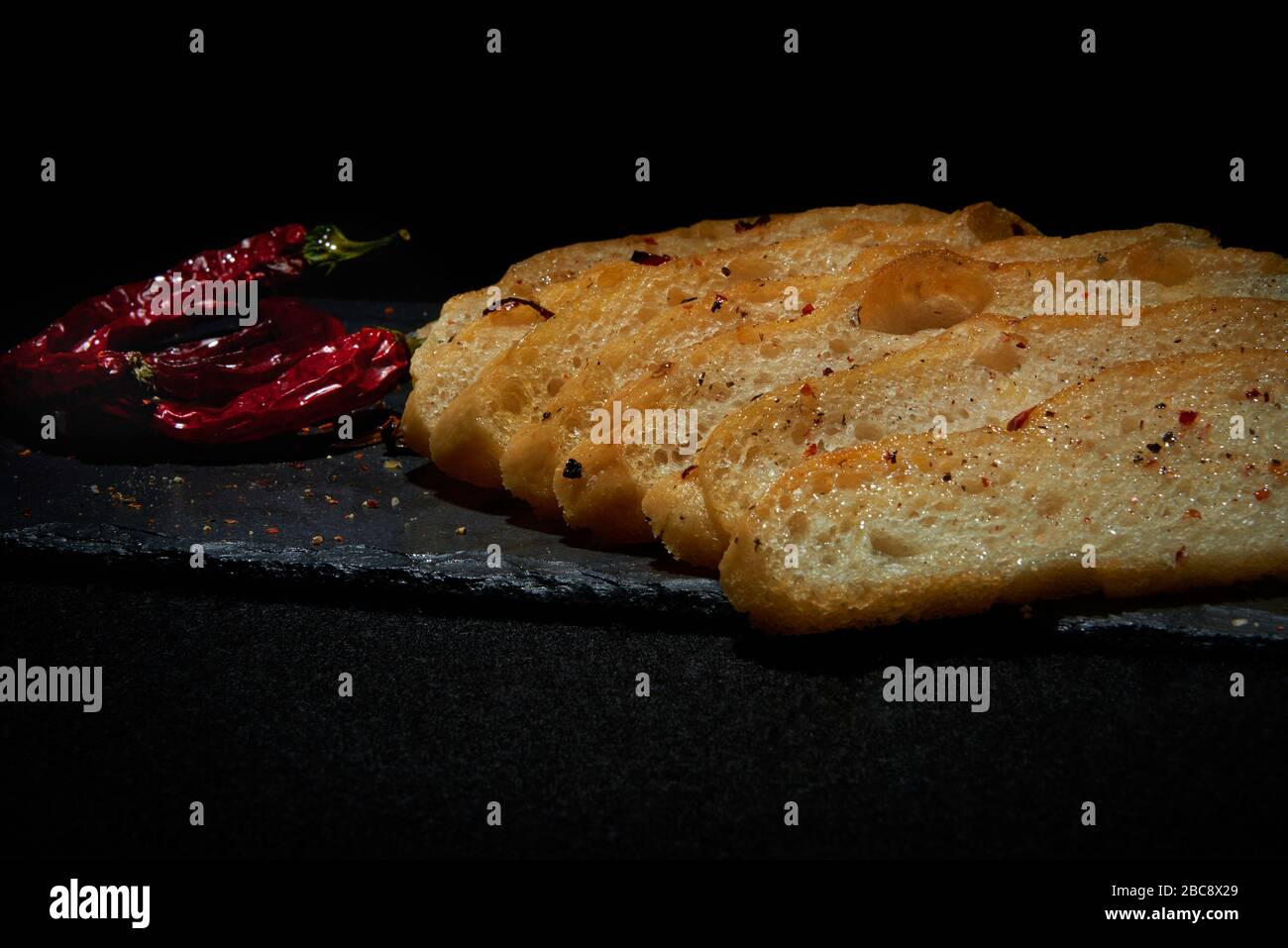 Display of homemade Italian style Focaccia loaf with dried chillies.  Sliced bread displayed on modern black slate with red chillies and flakes Stock Photo
