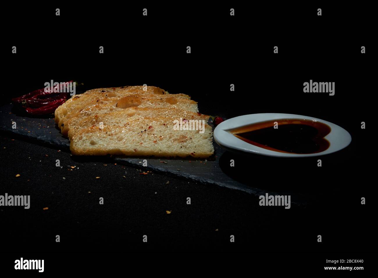 Display of homemade Italian style Focaccia bread with red chillies.  Sliced bread on black slate with balsamic vinegar in heart shape dish a Stock Photo