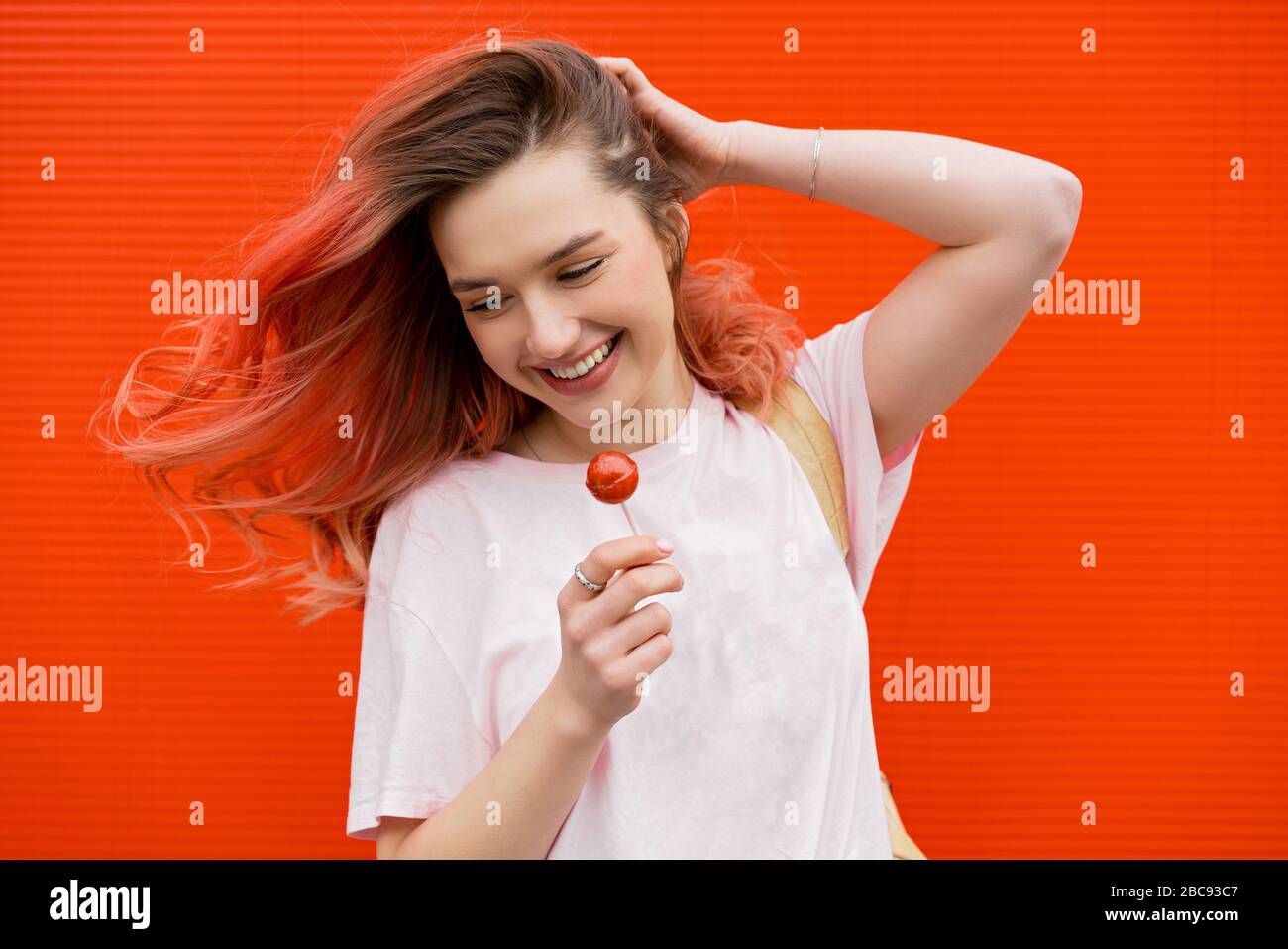 Fashion portrait of pretty smiling hipster woman with red flying haies and lollipop against the colorful orange wall Stock Photo