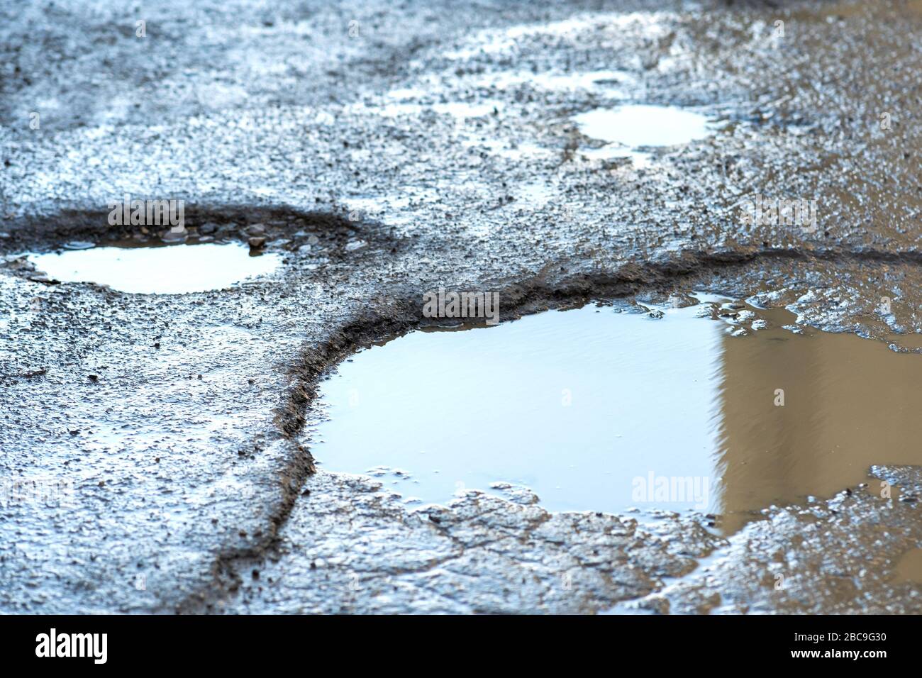 Close up of a road in very bad condition with big potholes full of dirty rain water pools Stock