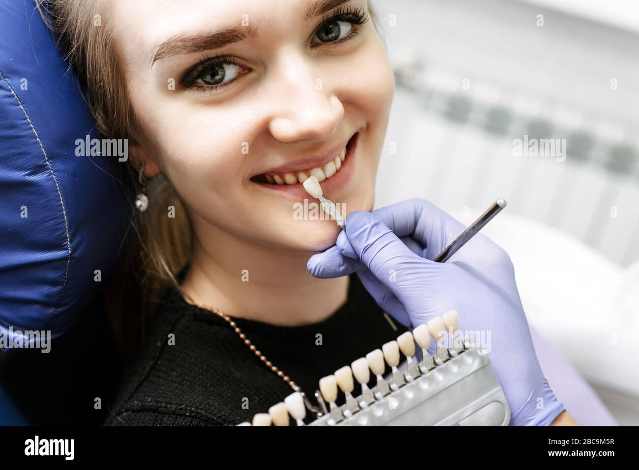 Dentist checking on female patient teeth color, using tooth enamel scale, dental office interior. The blonde smiles in the dental chair, looking forward to the whitening procedure. Stock Photo