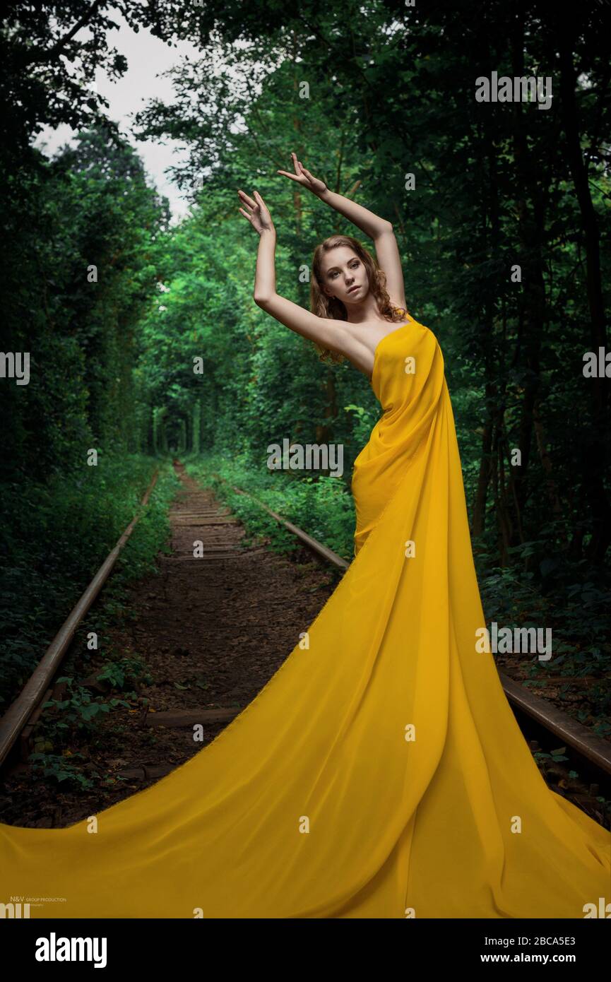 Beautiful young woman standing in a long yellow dress in nature tunnel Stock Photo