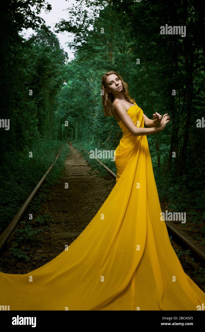 Beautiful young woman standing in a long yellow dress in nature tunnel Stock Photo