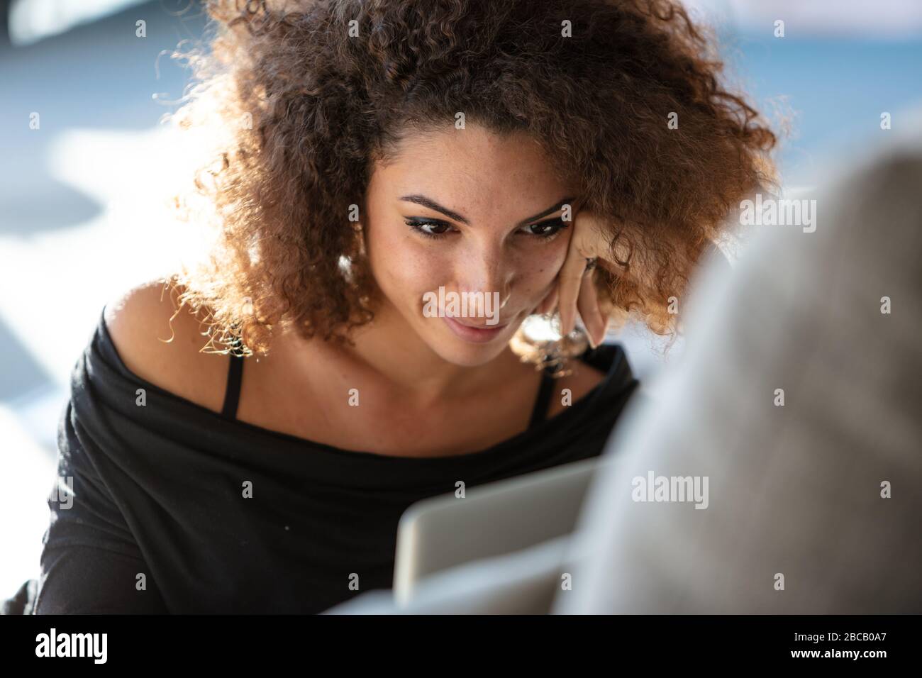 Close-up portrait of a beautiful smiling young woman in black dress and with curly brown hair, propping his face with her hand and looking at laptop s Stock Photo