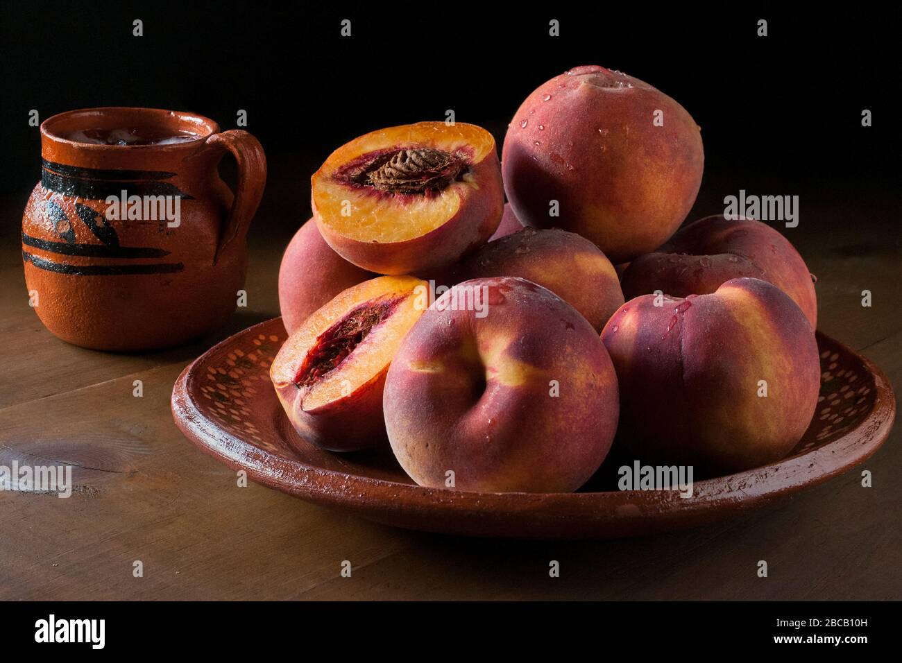 Peaches on a clay or mud Mexican plate with a Mexican mud jar with water in it, in dark food photography or low key light, macro photography. Stock Photo