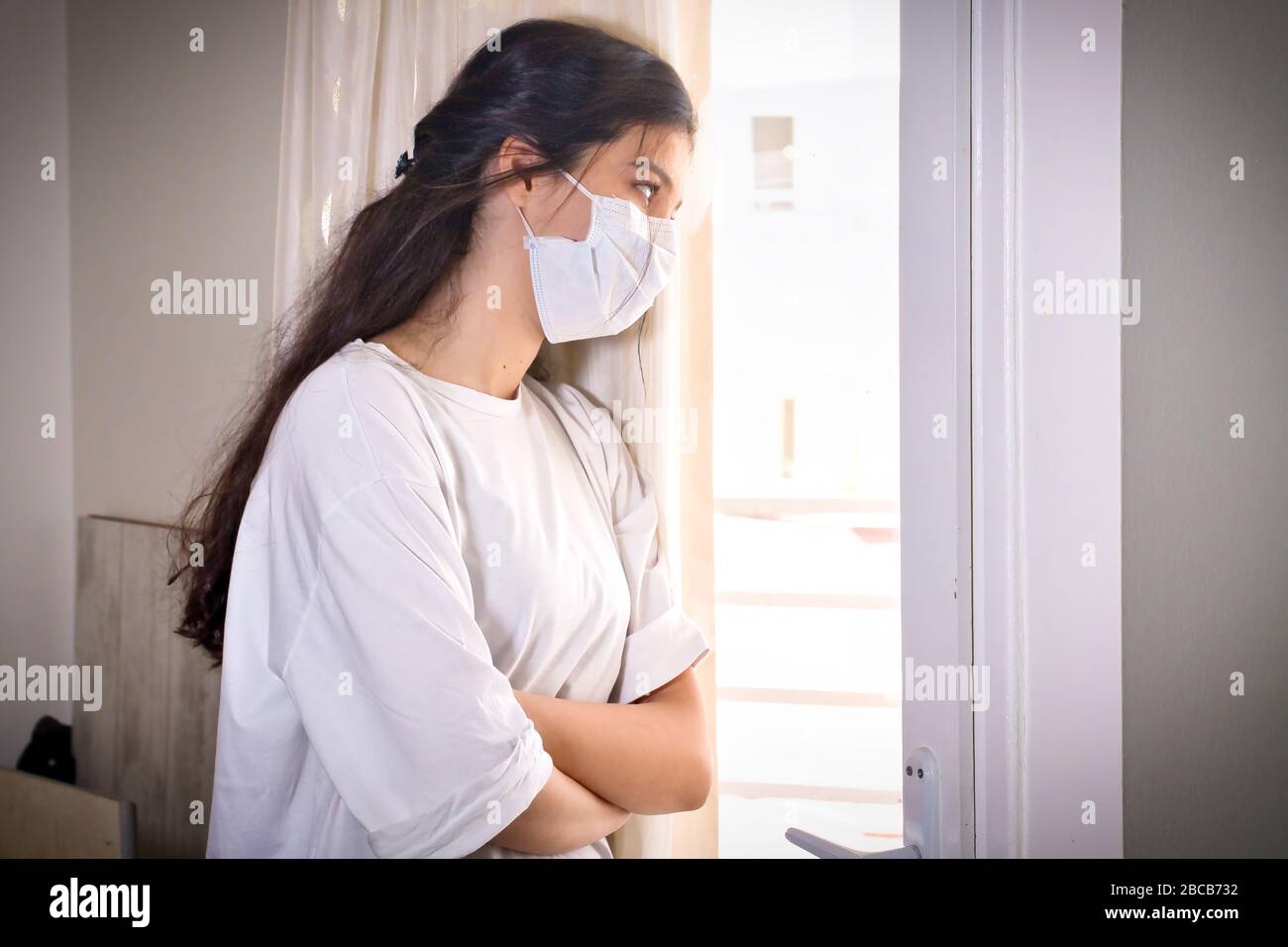 Asian woman doctor wearing a medical mask is looking out of the window with longing. The coronavirus spends quarantine days at home. Stock Photo