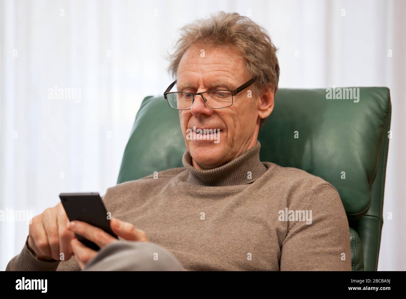 Senior man sitting in a chair in a bright living room looking at a cell phone at home Stock Photo