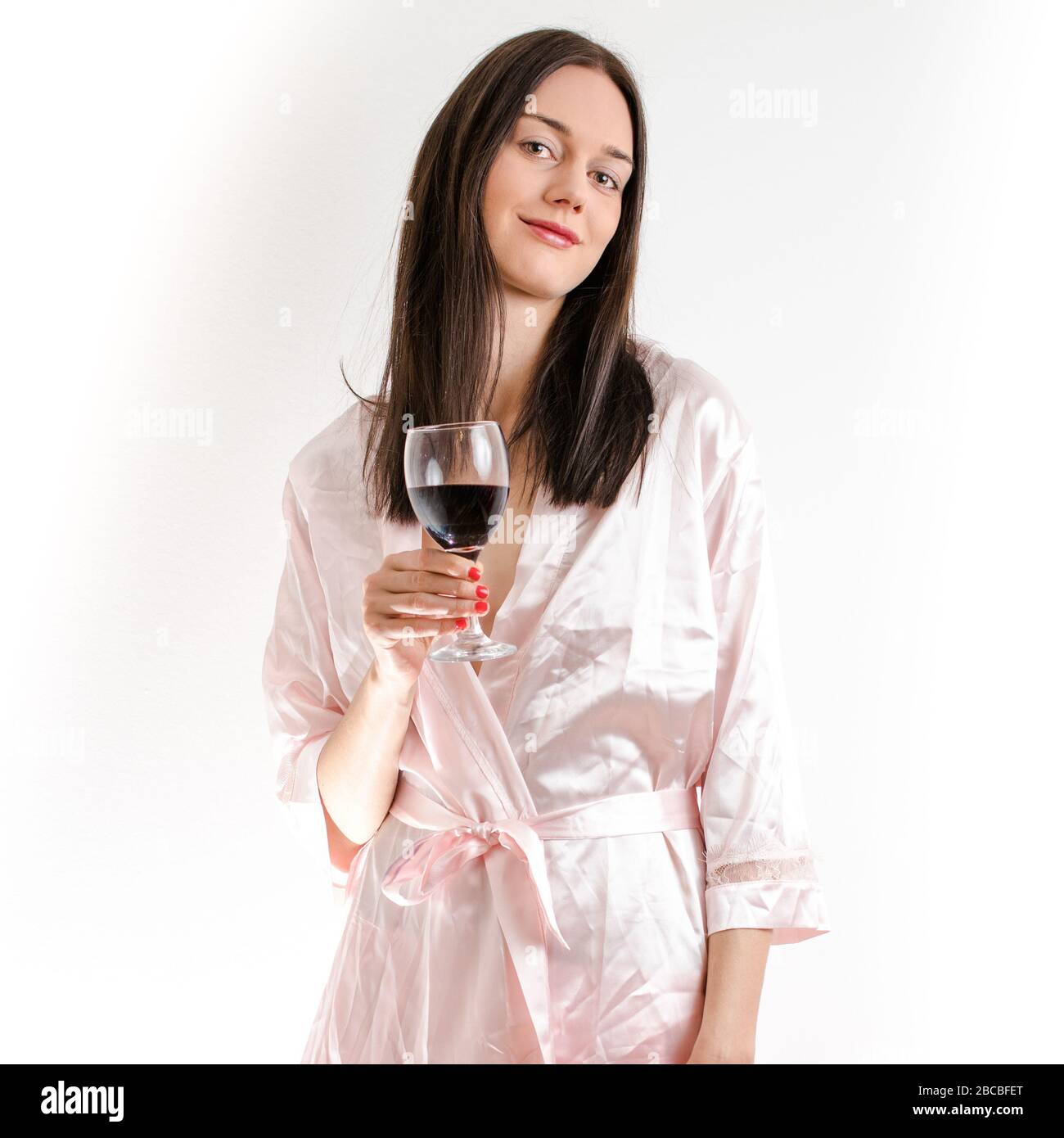 Attractive young brunette woman wearing a pink nightgown, holding a class of red wine smiling. Royalty free stock photo. Stock Photo