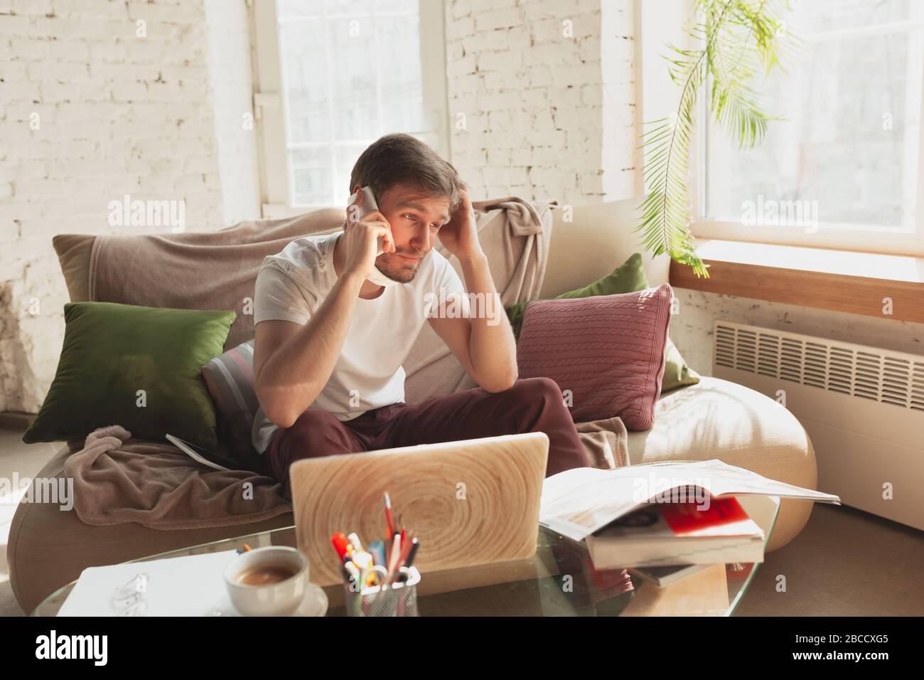 Young man studying at home during online courses for designers, SMM, SEO, analytics. Getting profession while isolated, quarantine against coronavirus spreading. Using laptop, smartphone, headphones. Stock Photo