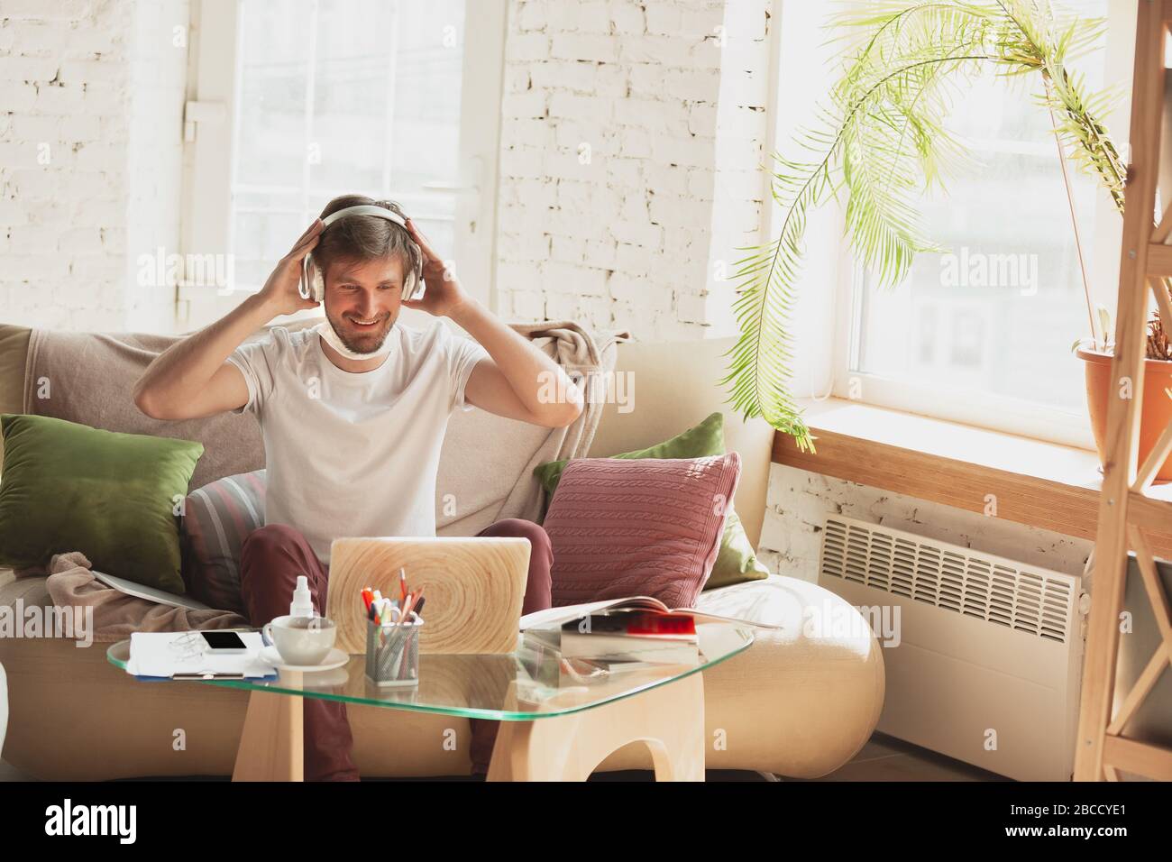 Young man studying at home during online courses for marketer, architect, translator. Getting profession while isolated, quarantine against coronavirus spreading. Using laptop, smartphone, headphones. Stock Photo