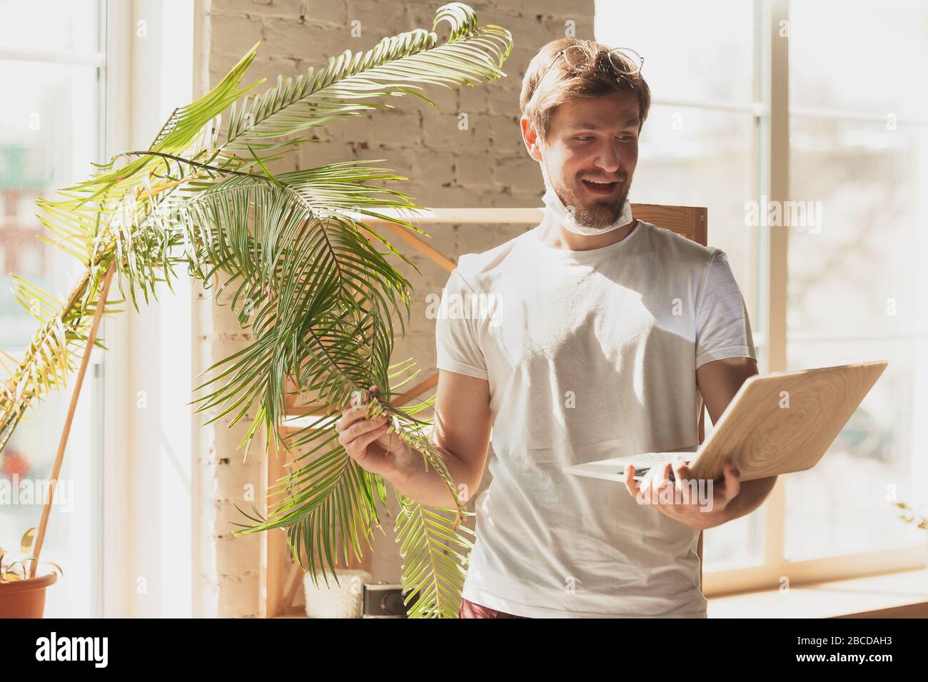 Young man studying at home during online courses for gardener, biologist, florist. Getting profession while isolated, quarantine against coronavirus spreading. Using laptop, smartphone, headphones. Stock Photo