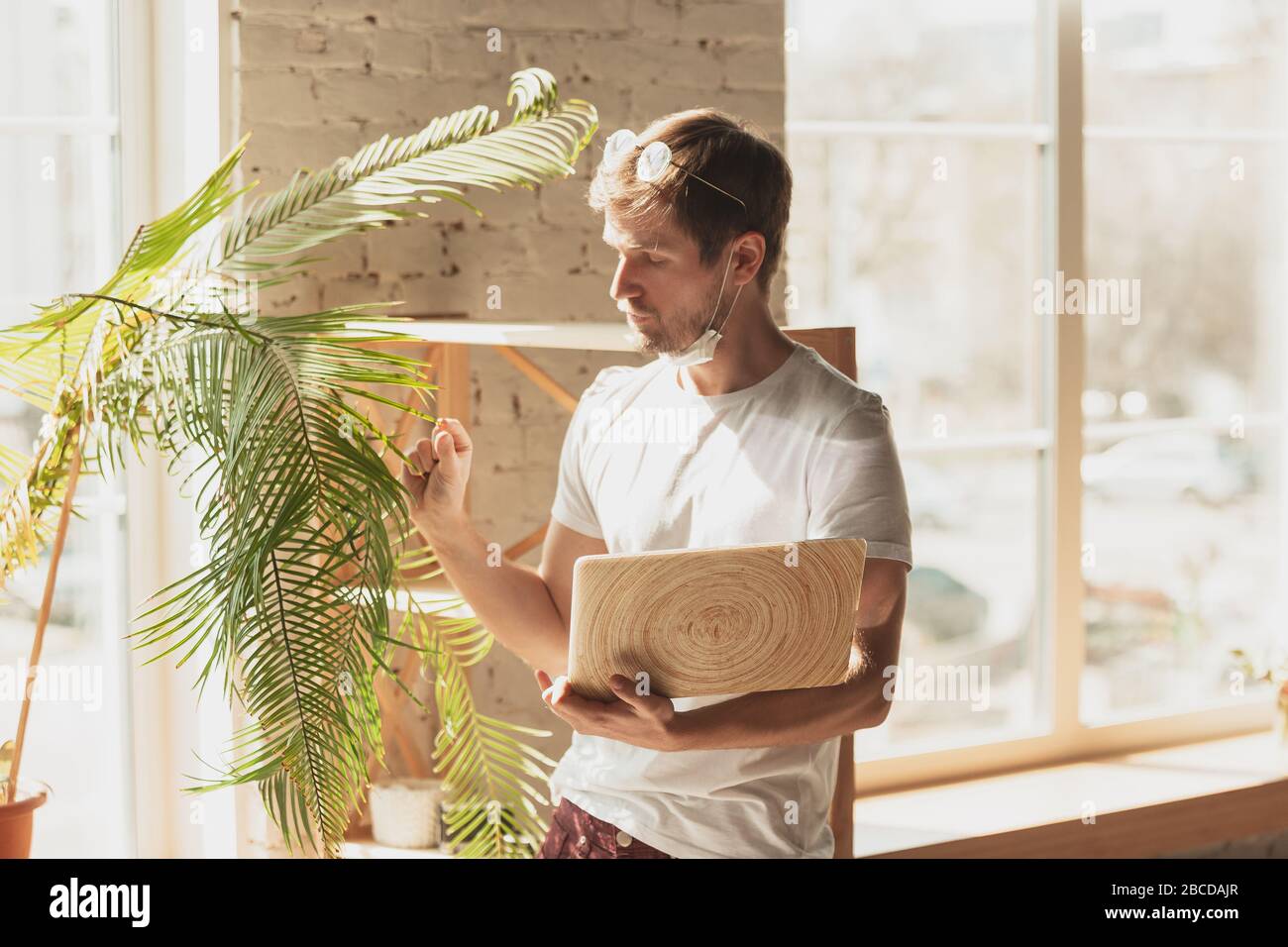 Young man studying at home during online courses for gardener, biologist, florist. Getting profession while isolated, quarantine against coronavirus spreading. Using laptop, smartphone, headphones. Stock Photo