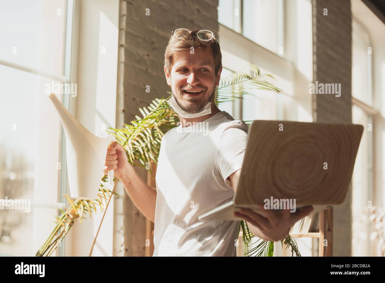 Young man studying at home during online courses for gardener, biologist, florist. Getting profession while isolated, quarantine against coronavirus spreading. Using laptop, smartphone, headphones. Stock Photo