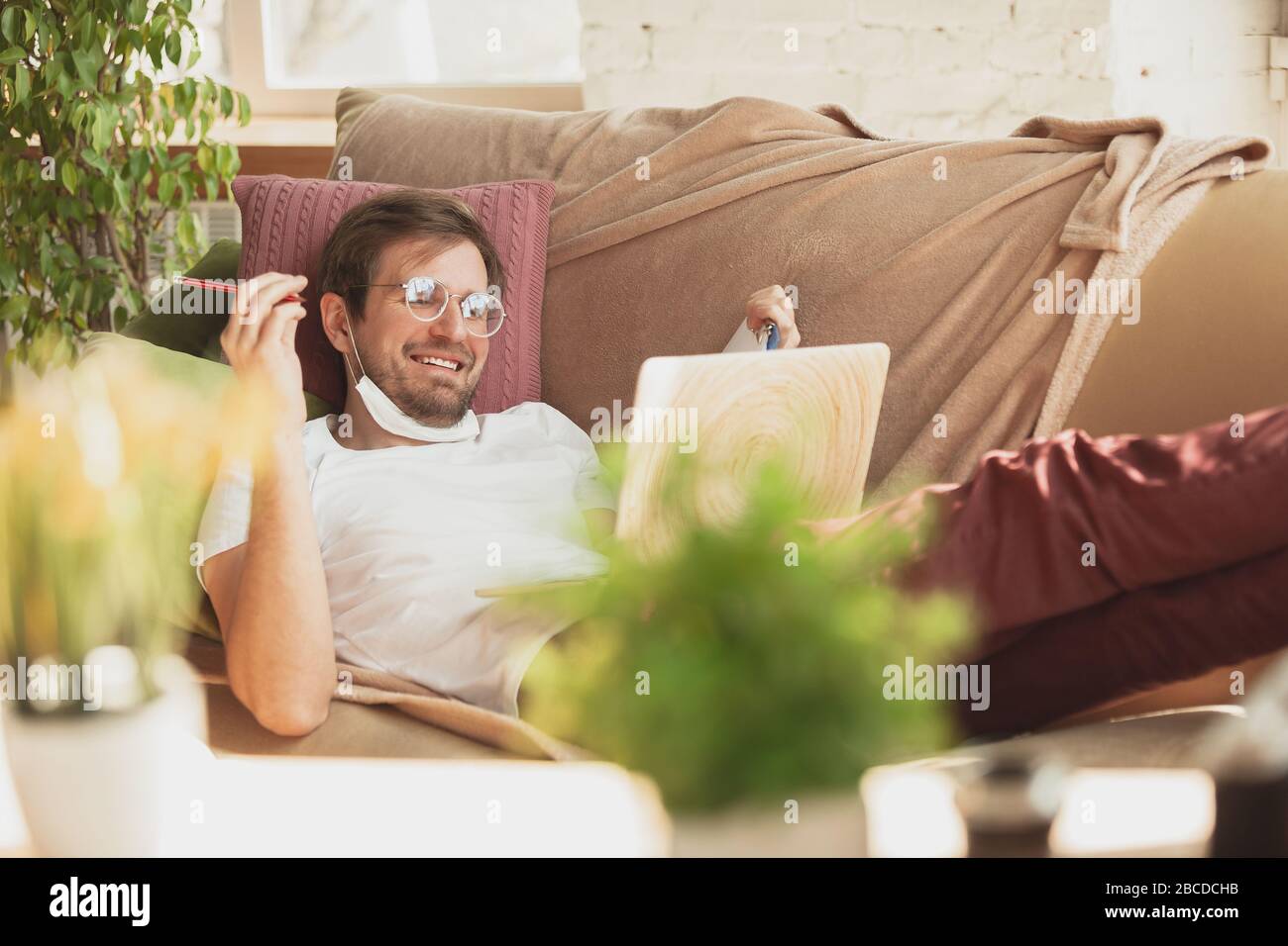 Young man studying at home during online courses for journalist, critics, writers. Getting profession while isolated, quarantine against coronavirus spreading. Using laptop, smartphone, headphones. Stock Photo