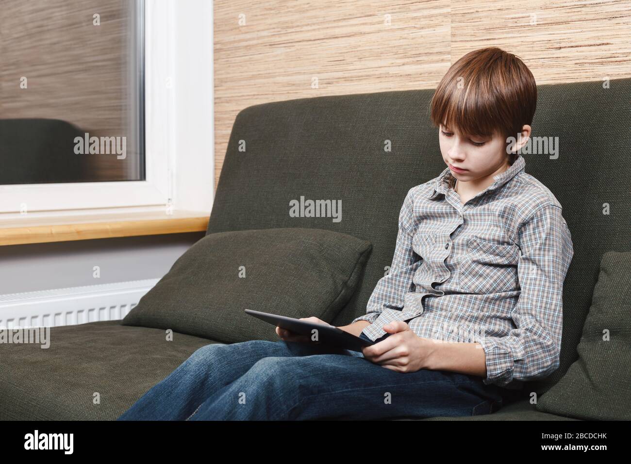 Lonely boy sitting on sofa with tablet computer stuck at home during