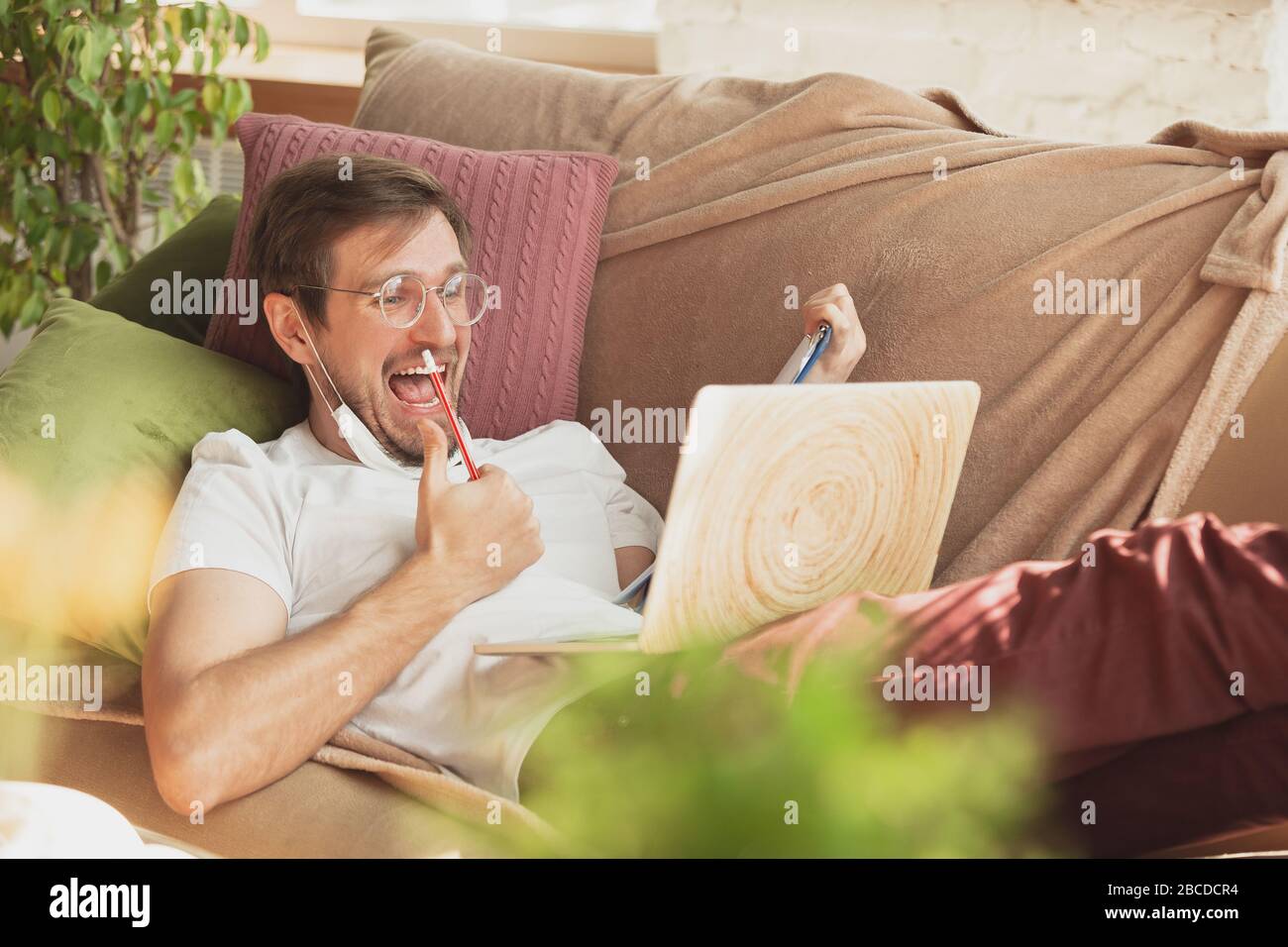 Young man studying at home during online courses for journalist, critics, writers. Getting profession while isolated, quarantine against coronavirus spreading. Using laptop, smartphone, headphones. Stock Photo