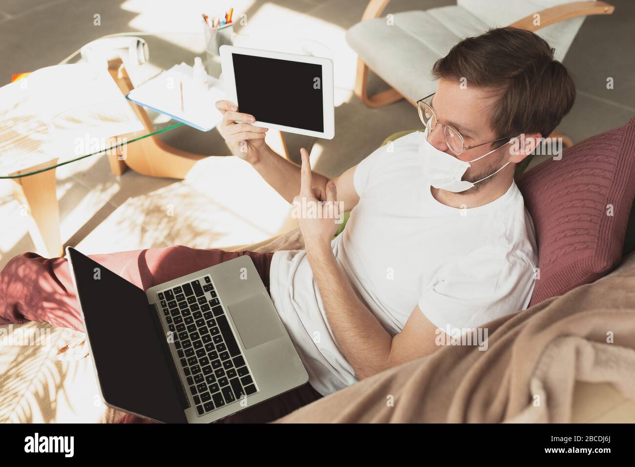 Young man studying at home during online courses for disinfector, nurse, medical services. Getting profession while isolated, quarantine against coronavirus. Using laptop, smartphone, headphones. Stock Photo