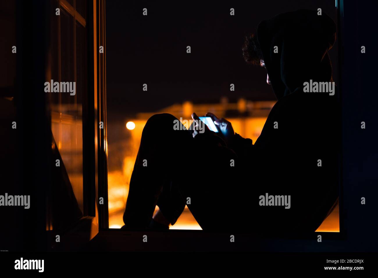 Young man with mobile phone confined to his house watching out the window at night. Stock Photo