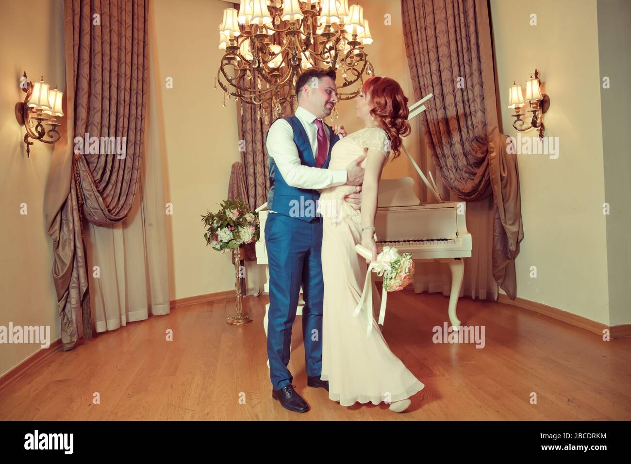 Bridal couple in front of a piano . Happy wedding day. Happy bride and groom near piano in white interior studio . Newlyweds stand in the interior Stock Photo