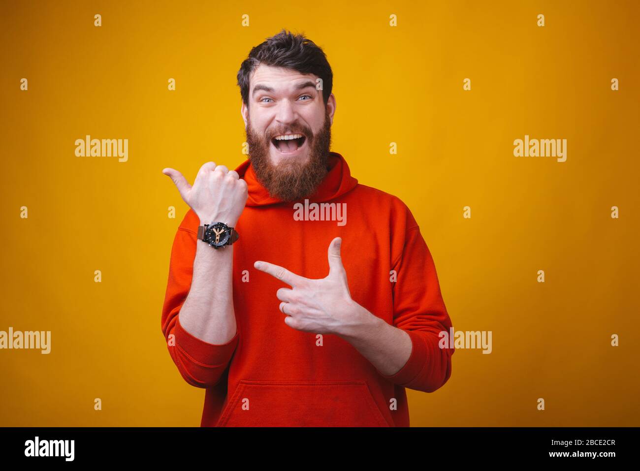 Photo of surprised bearded man is pointing at his wrist watch on yellow background. Its time. Stock Photo