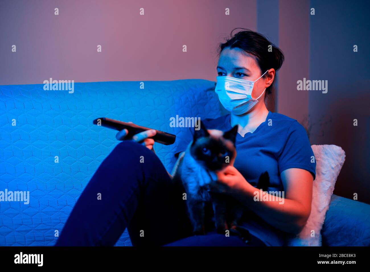 Female with mask sitting on sofa near her cat and look at tv. Concept of persona and his pet in quarantine (self-isolation) Stock Photo