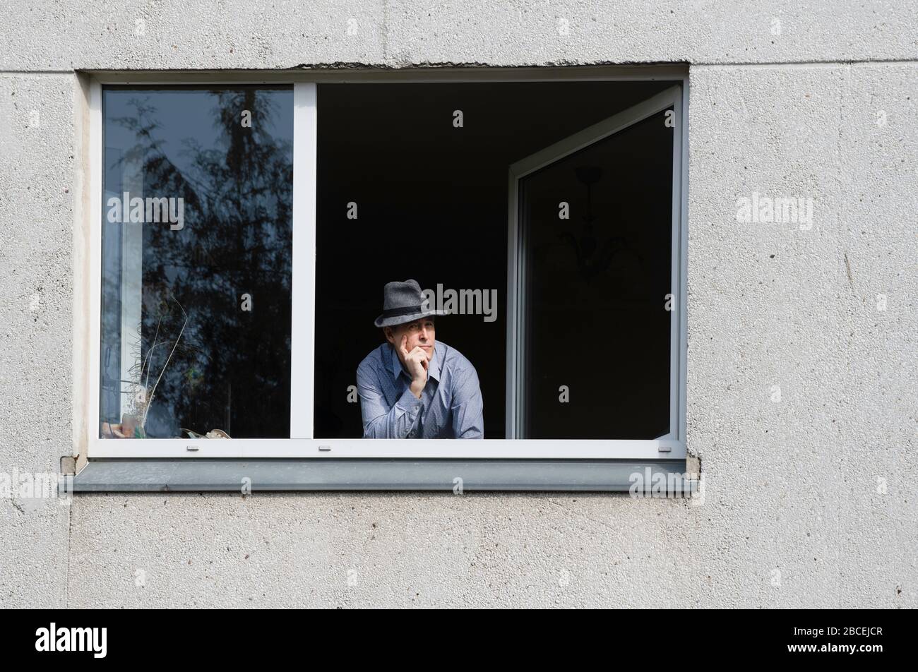 Bored man in corona quarantine looking out of window to the street.Man with the hat celebrating alone birthday at home during Covid-19 Lockdown. Sad b Stock Photo