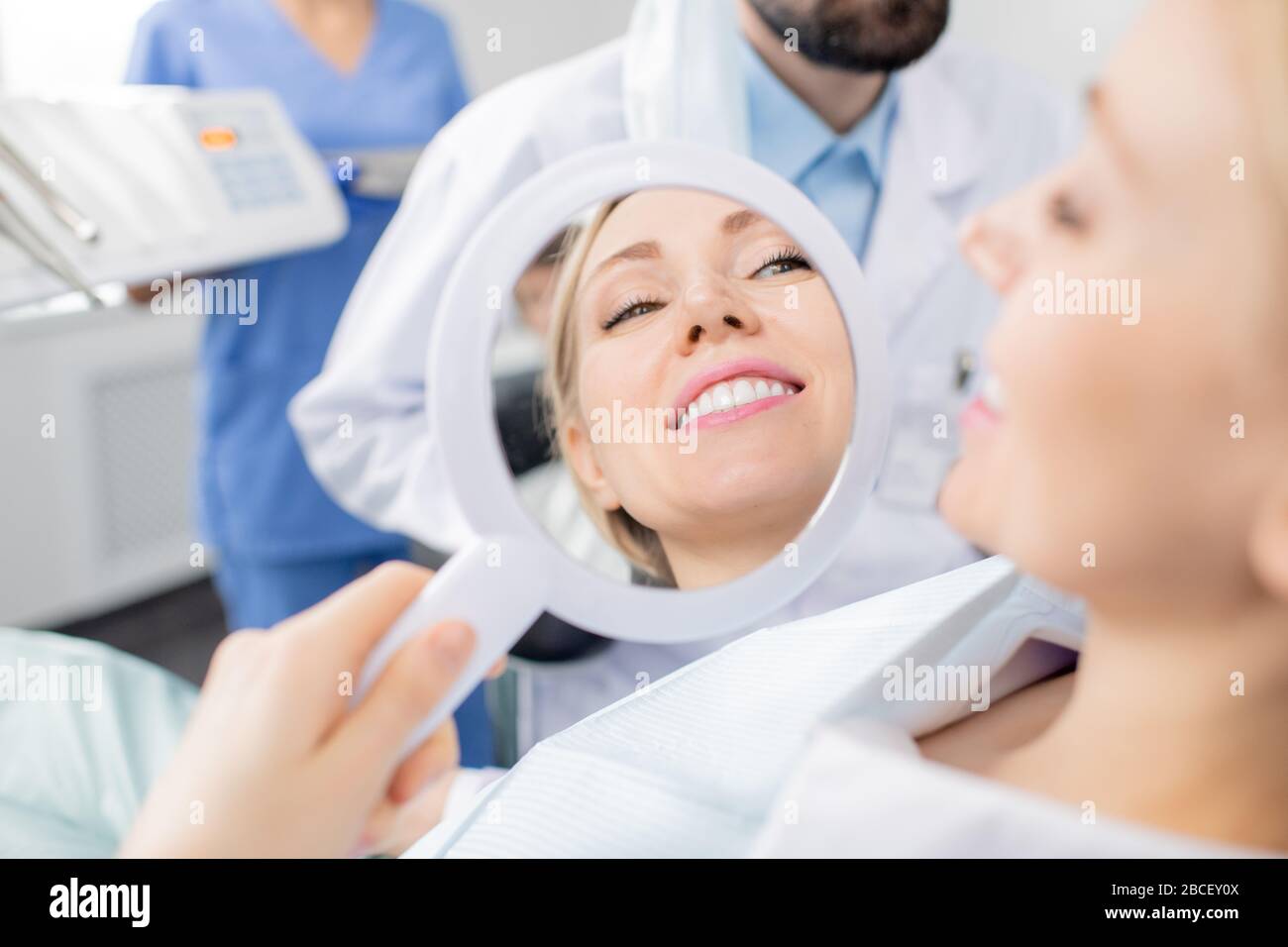 Reflection in mirror of healthy smile of pretty young smiling female patient of dental clinics after whitening teeth procedure by her dentist Stock Photo