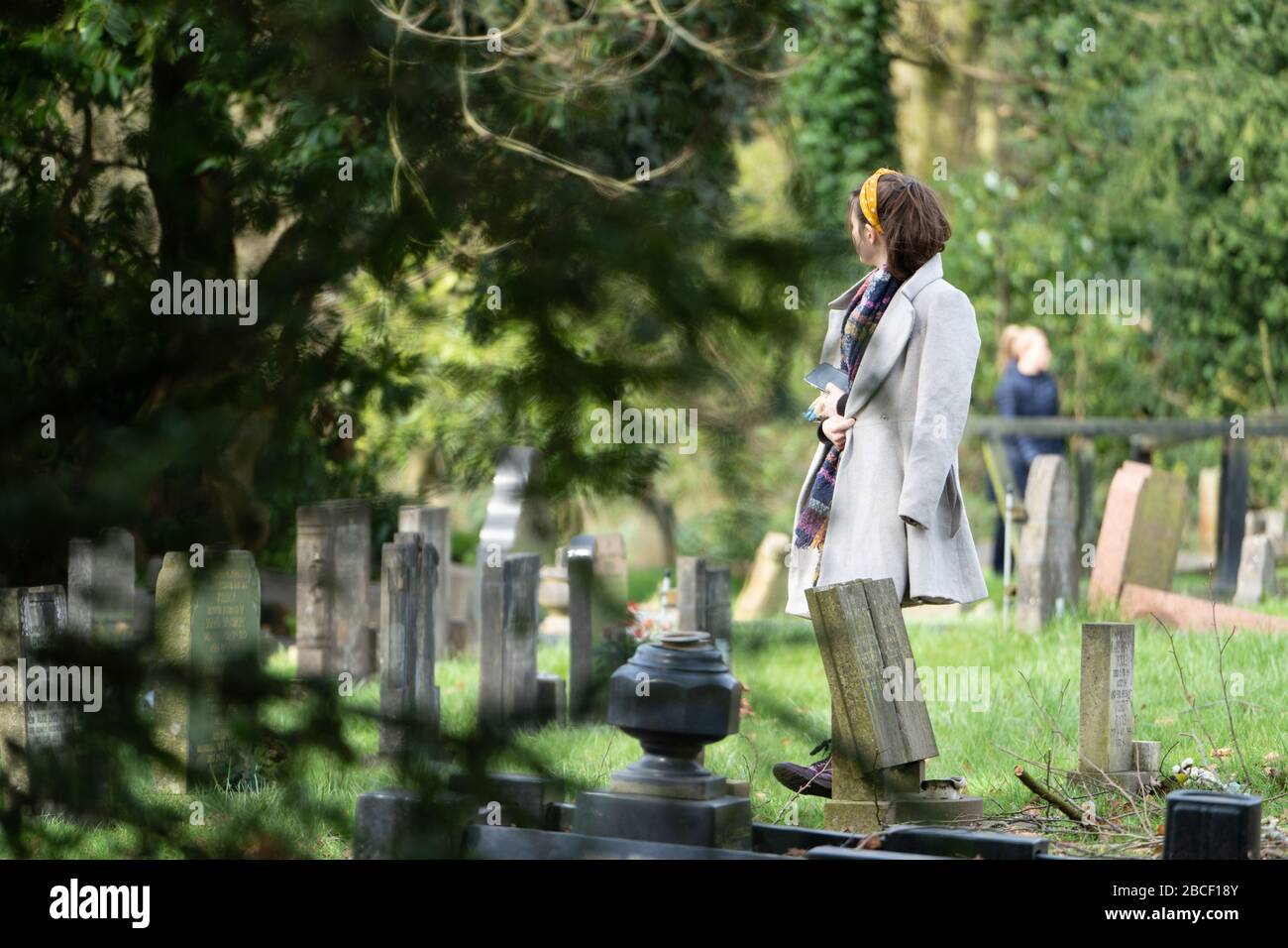 A young woman wearing a grey coat, thoughtfully standing amongst gravestones in a cemetery on a sunny day. UK Stock Photo