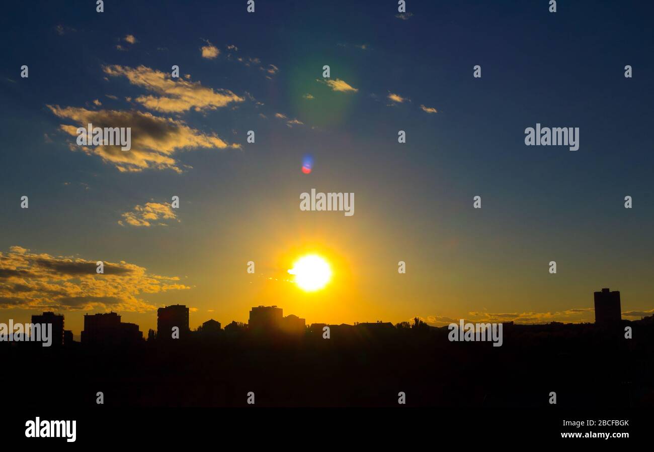 Lovely sunset, with golden sun, with gray-white clouds, with azure sky, with lens flares against the background of the city silhouette. Stock Photo