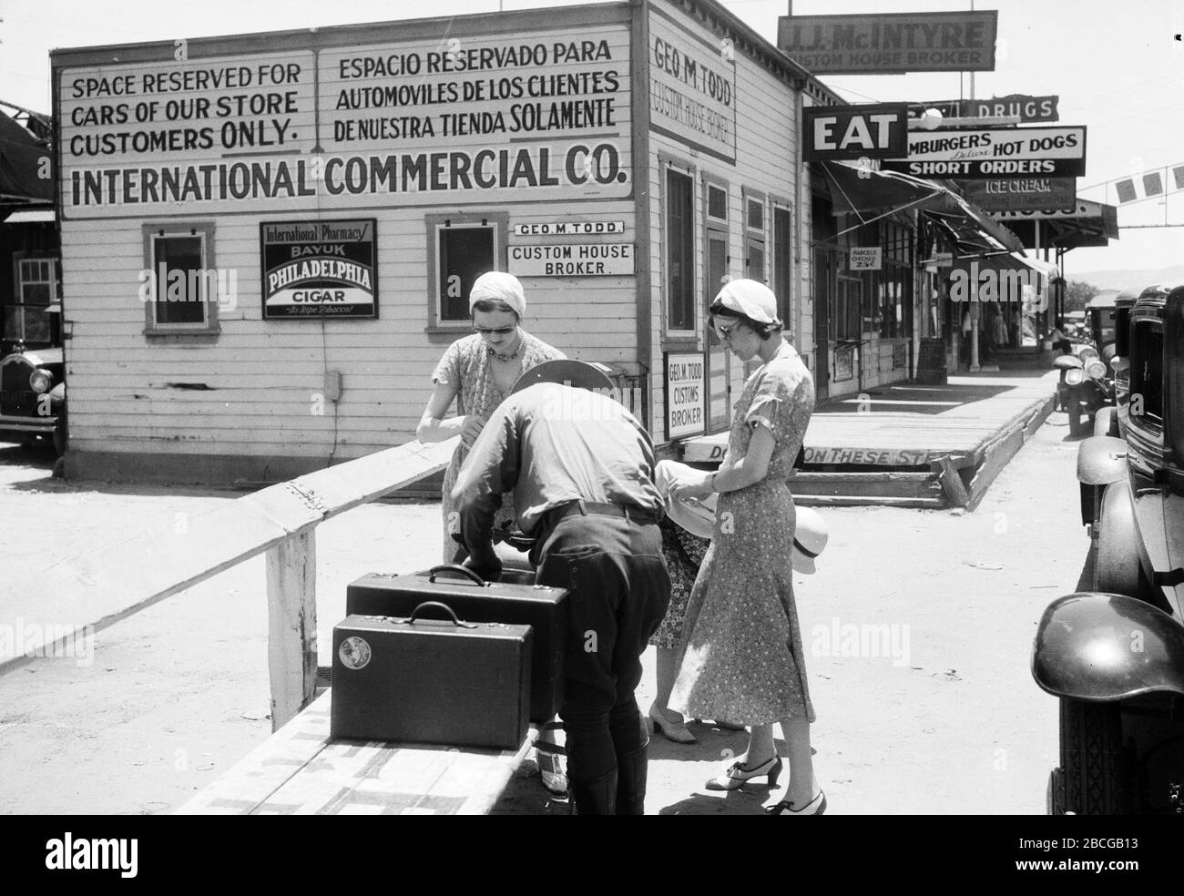 A male U.S. Customs Inspector examines baggage belonging to women at the United States - Mexico border at Tijuana, Mexico and San Ysidro, California, 1920s. Nearby are shops and offices for Customs