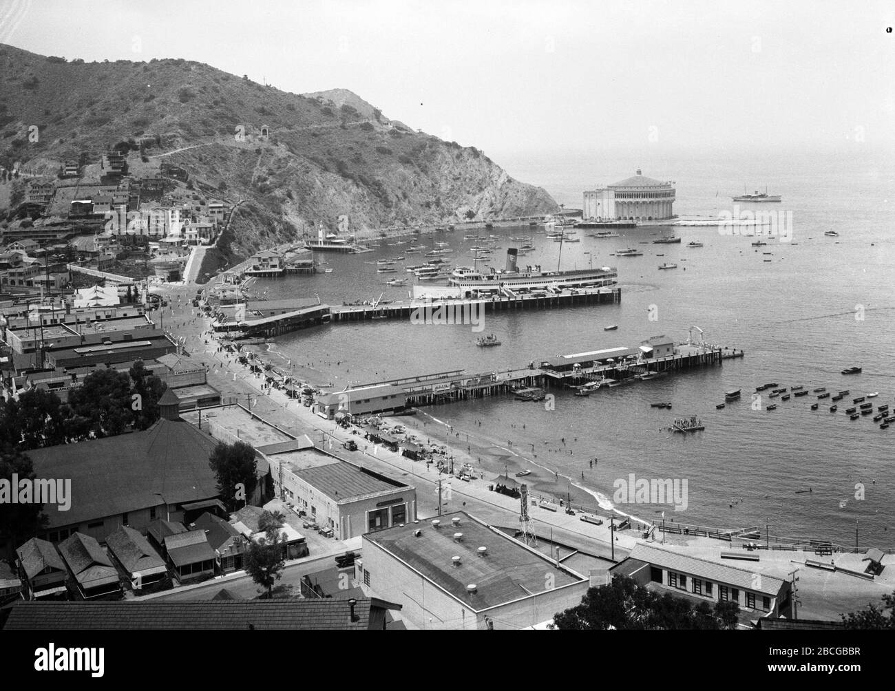 High angle view of Avalon Bay, Avalon, Santa Catalina Island, California, 1931. In the harbor boats are moored and a steamship docked at a pier, people line the beaches and wade in the water, and the waterfront road leads to the Catalina Casino Ballroom. Photography by Burton Holmes. Stock Photo