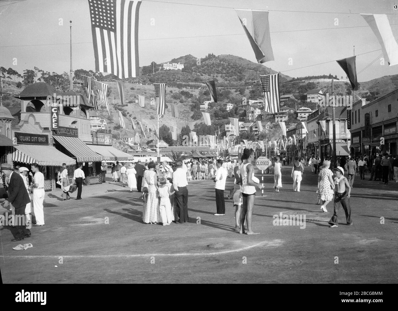 Pedestrians wander around a wide street closed to vehicle traffic, possibly Crescent Avenue, in Avalon, Santa Catalina Island, California, 1931. Cafés and storefronts are seen to the left and right, flags are suspended overhead and hillside houses are seen in the distance. Photography by Burton Holmes Stock Photo