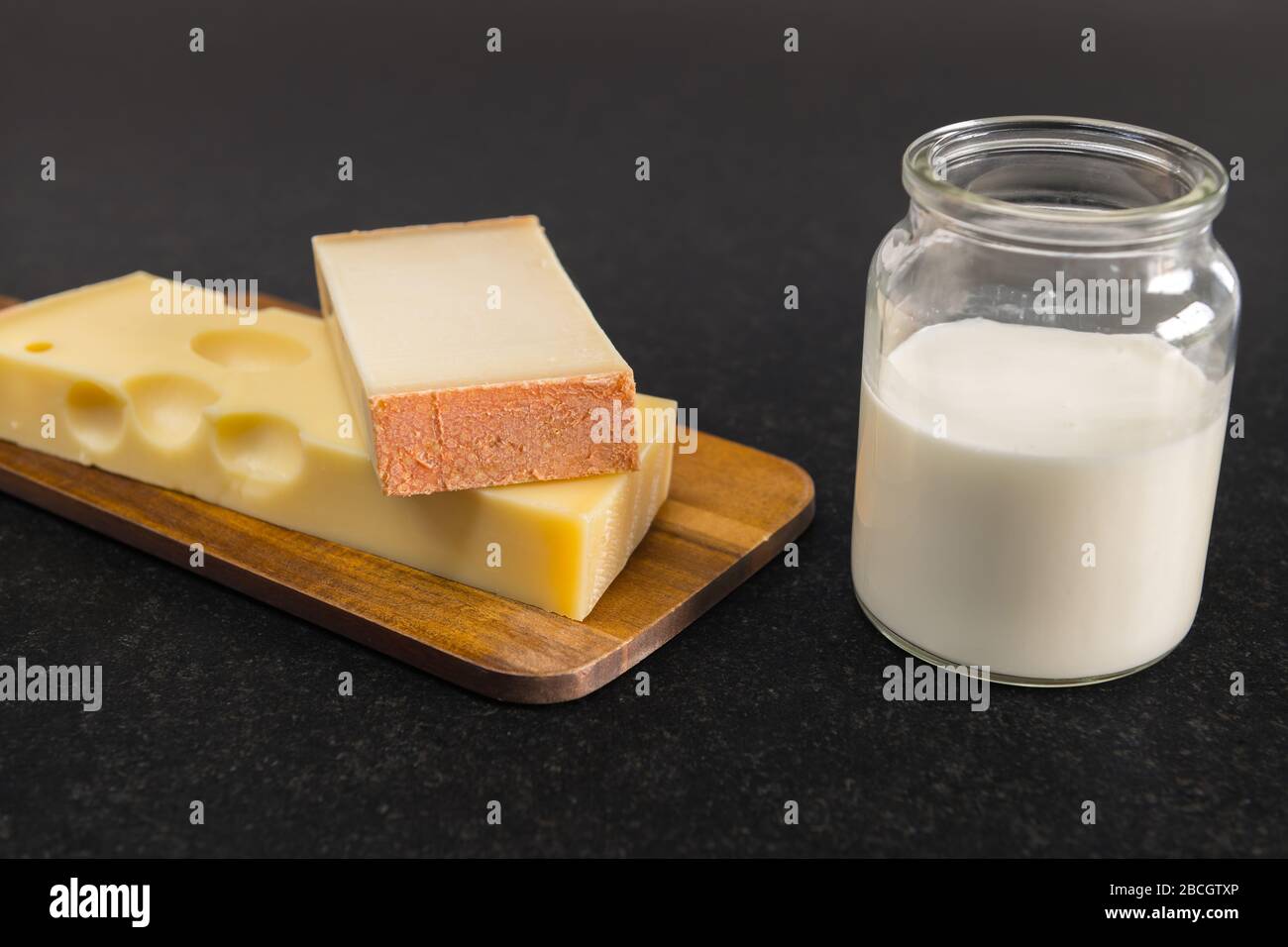 Emmental cheese and Gruyere cheese on a wooden board stand next to a glass filled with milk on a black table. Stock Photo