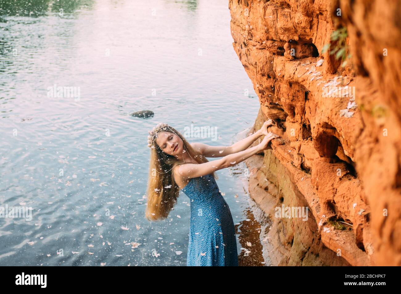 Young woman with closed eyes in crown from shells and threw back head staying near red sand wall in river Stock Photo