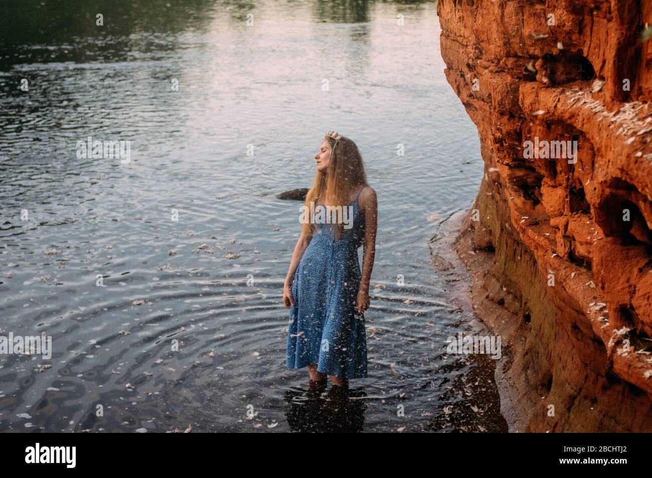 Young woman with crown from shells and threw back head staying near red sand wall in river, closed eyes Stock Photo