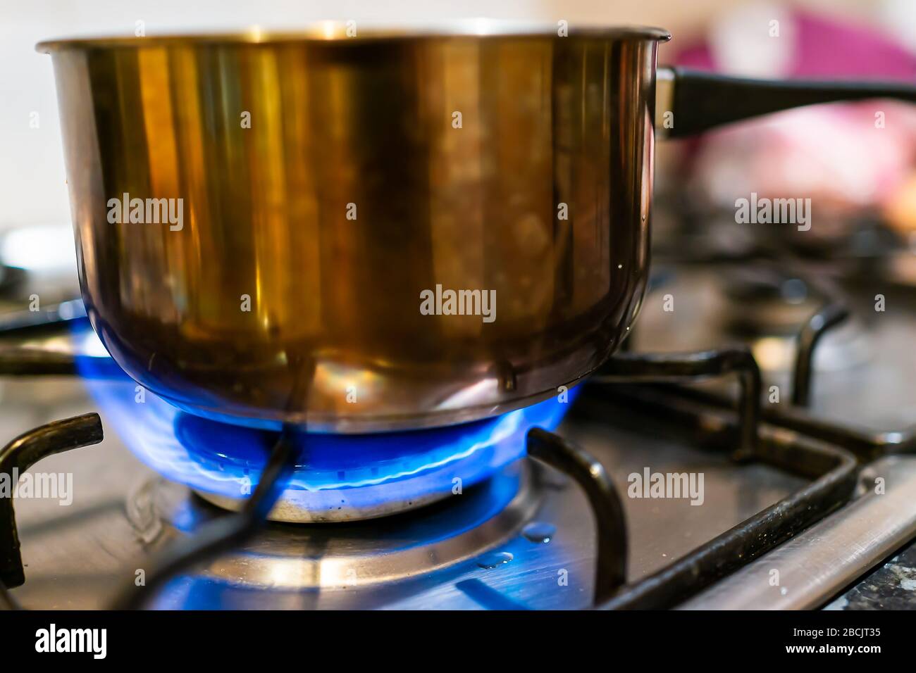 Macro closeup of gas stove top with blue fire flame and stainless steel pot with reflection and blurry blurred background Stock Photo