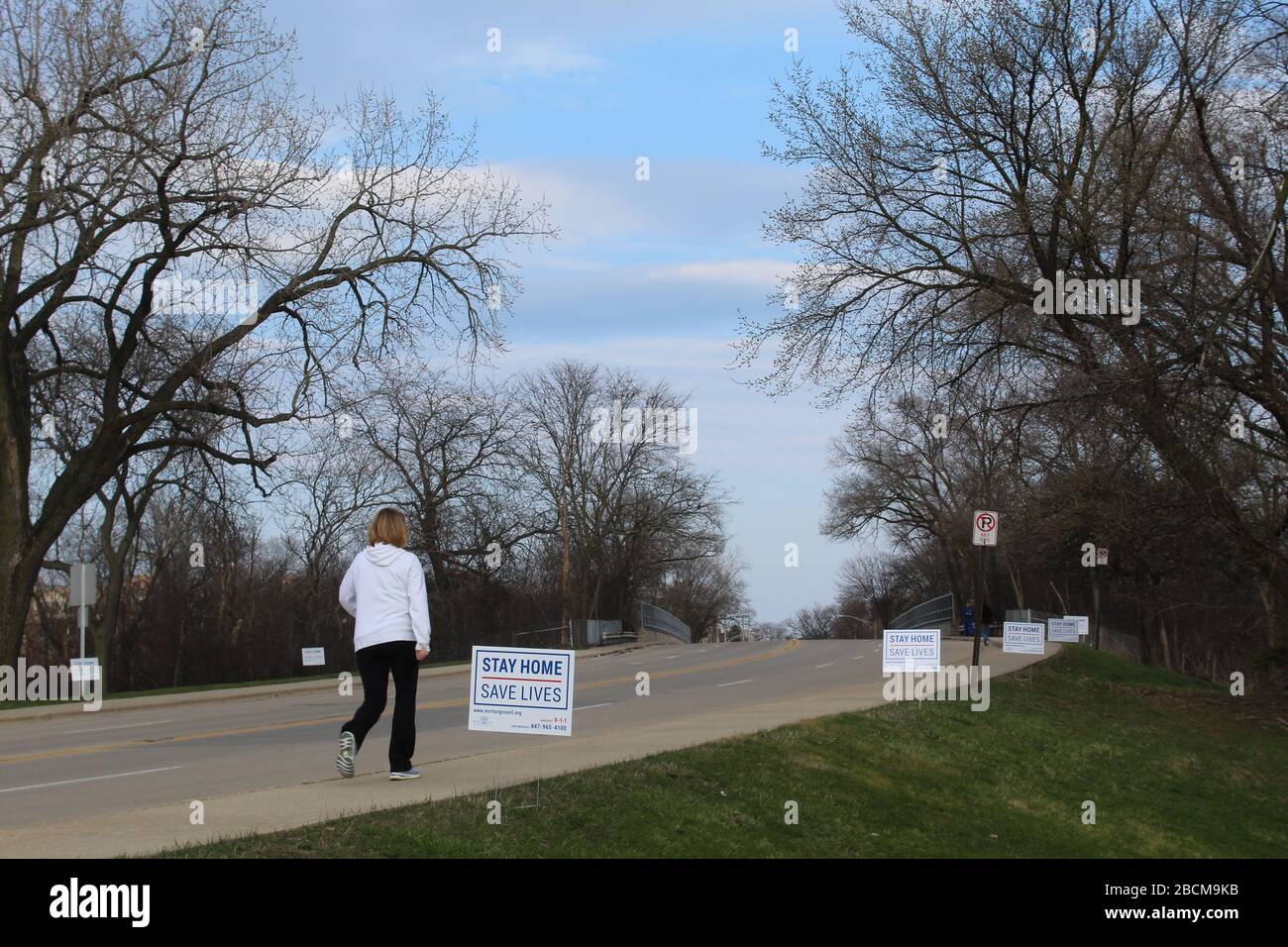 Woman walking uphill towards Stay Home Stay Lives signs during coronavirus pandemic shelter-in-place order Stock Photo