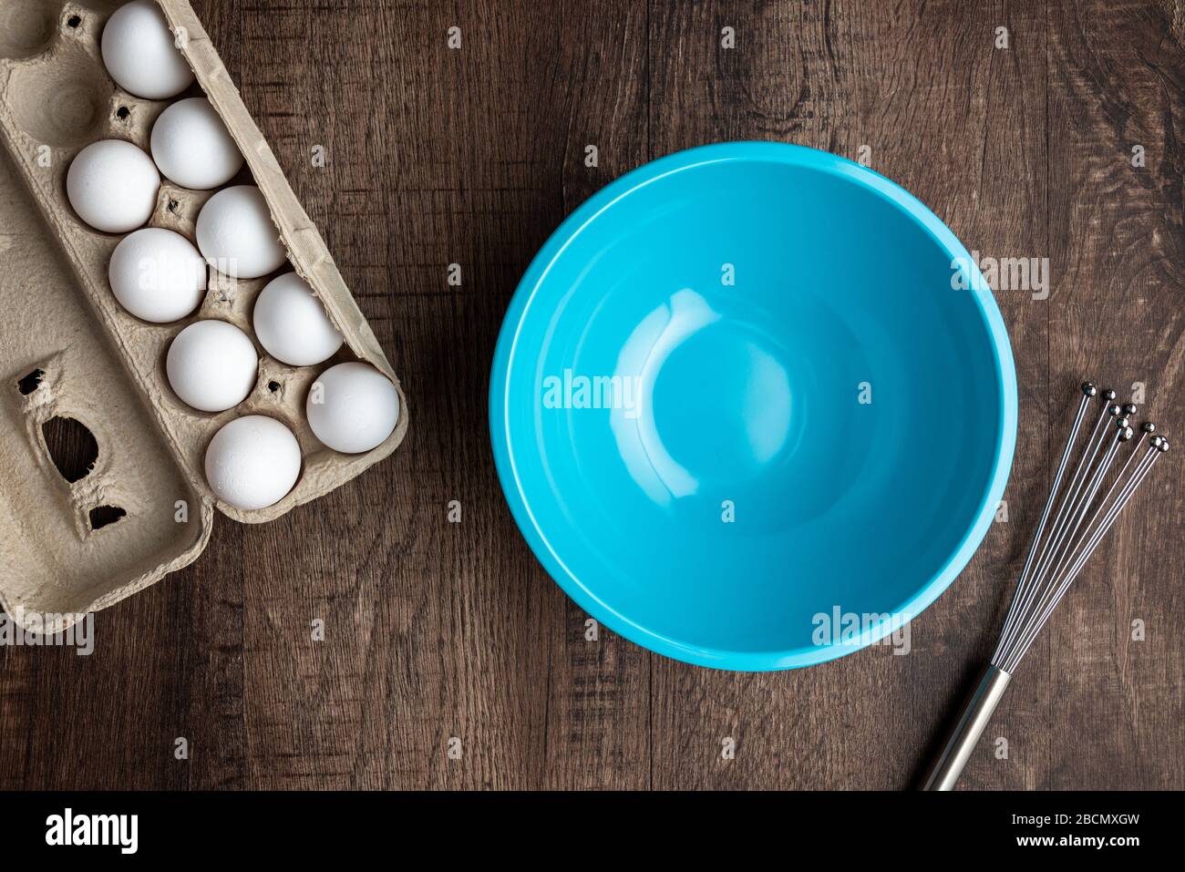 Whole eggs in cardboard egg carton, empty blue mixing bowl, metal whisk