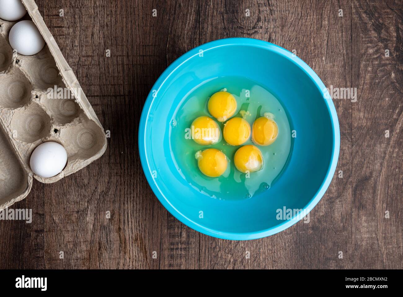 Six cracked eggs in blue mixing bowl, whole eggs in cardboard carton