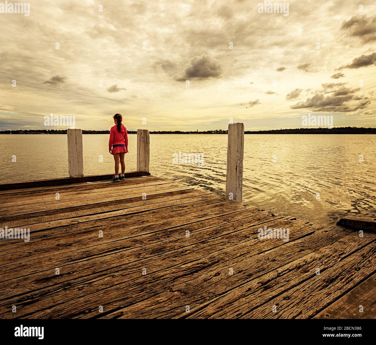 Young girl standing at the edge of a wooden jetty looking out into the ...