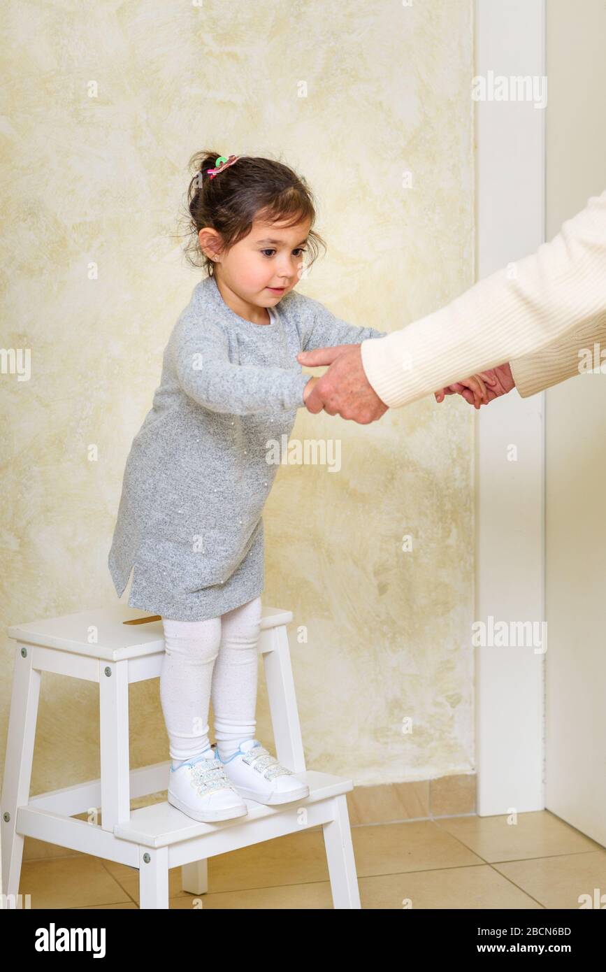 Beautiful little girl jumps off stepladder, holding hands with grandfather. Childish activity ideas, home games for kids and childhood concept. Stock Photo