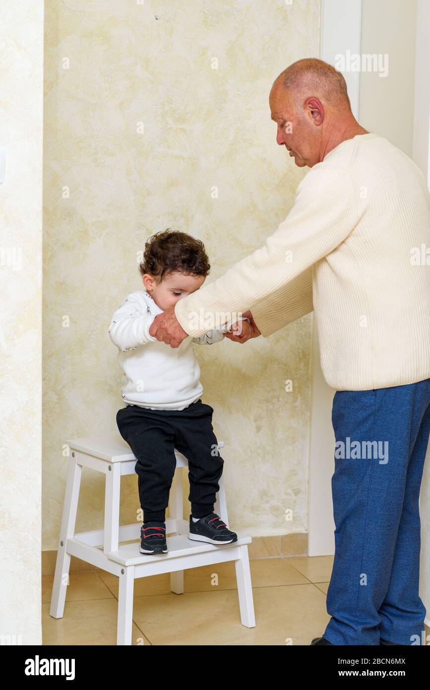Funny little boy jumps off stepladder, holding hands with grandfather. Childish activity ideas, home games for kids and childhood concept. Stock Photo