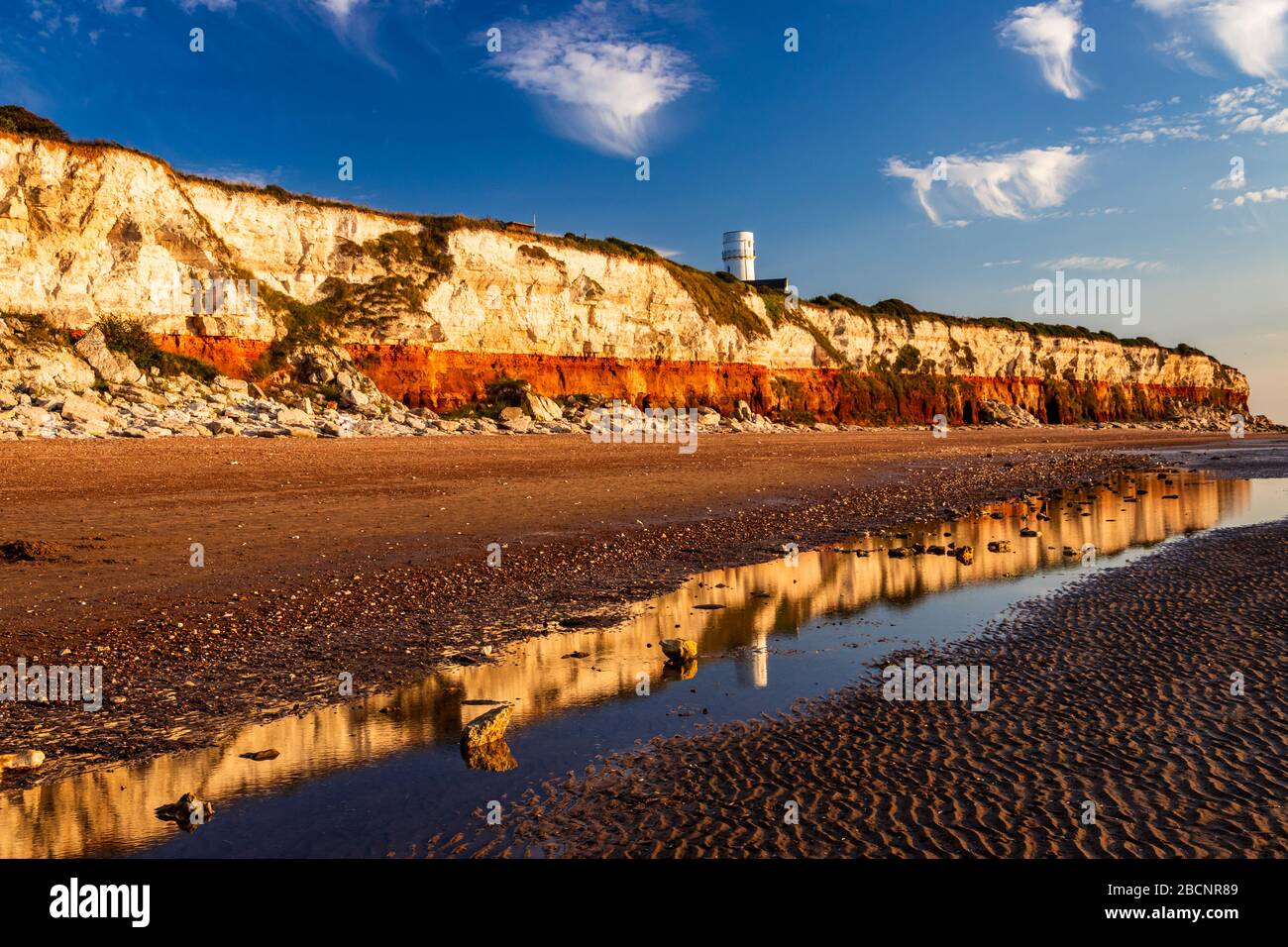Sun setting on Hunstanton beach in north Norfolk east Anglia illuminating the colourful cliff face and lighthouse reflecting in the low tide pools Stock Photo