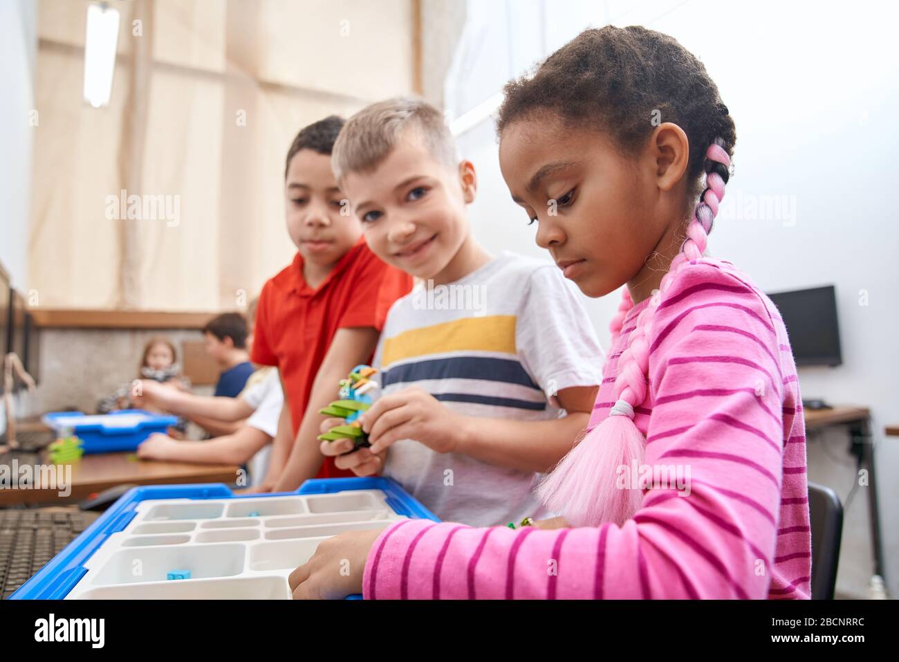 Side view of building kit with colorful peices in box for group of three multiracial kids creating toys. Close up of interested friends working on pro Stock Photo