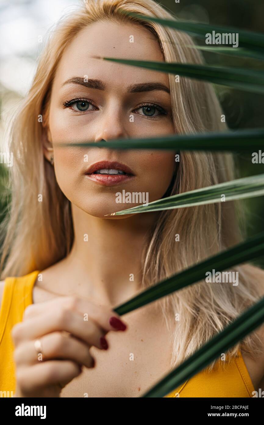 Close up beauty portrait of lovely caucasian blonde haired woman with clean skin, natural makeup, beautiful eyes, standing behind tropical leaf. Verti Stock Photo