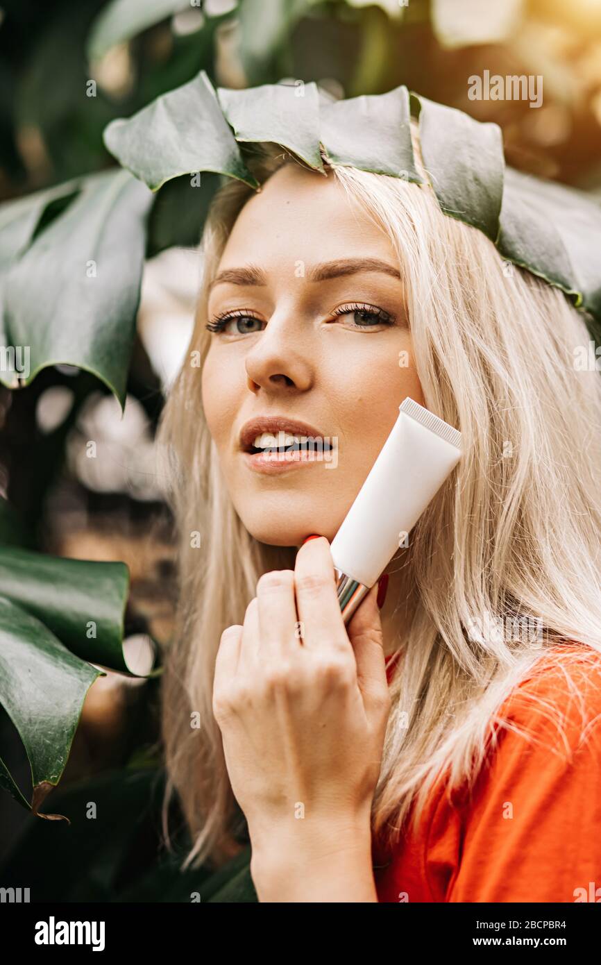 Gorgeous caucasian blonde woman with perfect clean skin, holding tube of cosmetic product, eye cream, sunscreen, standing on tropical plants backgroun Stock Photo