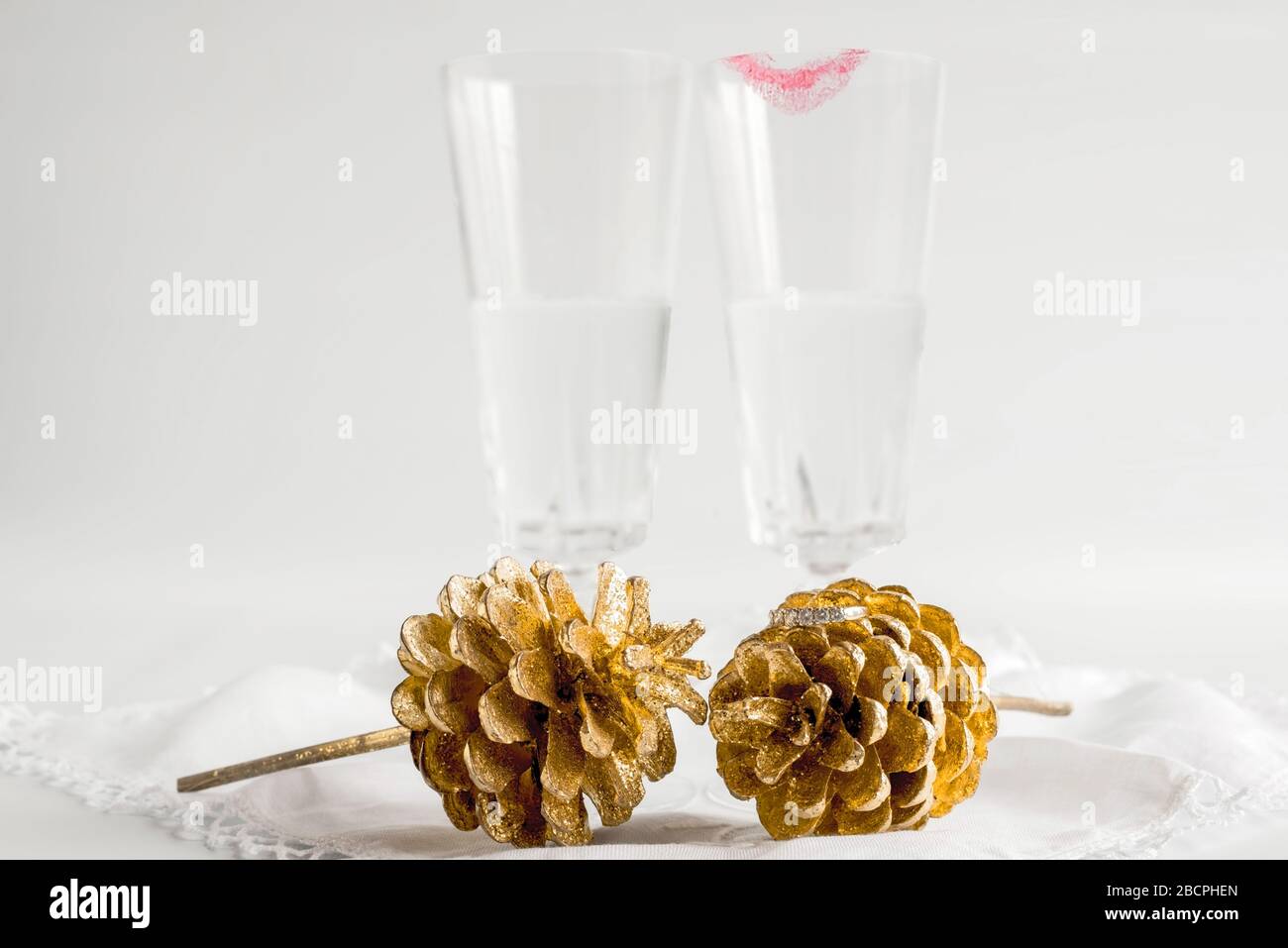 Two champagne glasses, one with lipstick, behind two gold fern cones. Diamond ring placed on one cone. Celebrating engagement, civil ceremony, wedding. Stock Photo