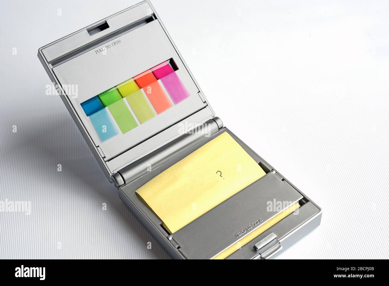 Silver dispenser of yellow postit notes and multi coloured sticky index highlighters against a white background, question mark written on postit note. Stock Photo